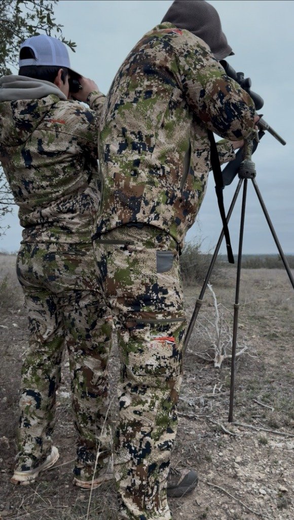Guide and hunter in camo outfits glassing distant terrain on tripod rifle mount