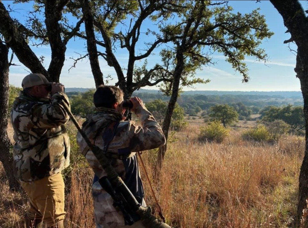 Two hunters with binoculars glassing expansive Texas Hill Country valley from oak ridge