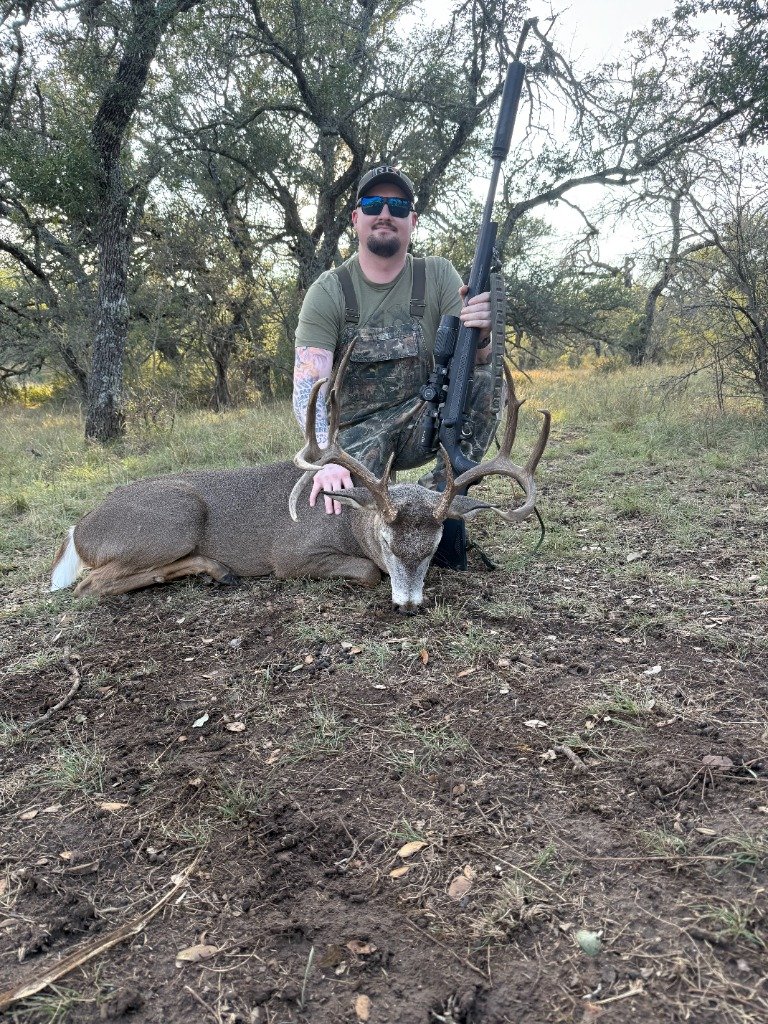Hunter posing behind wide heavy Whitetail buck in camo overalls